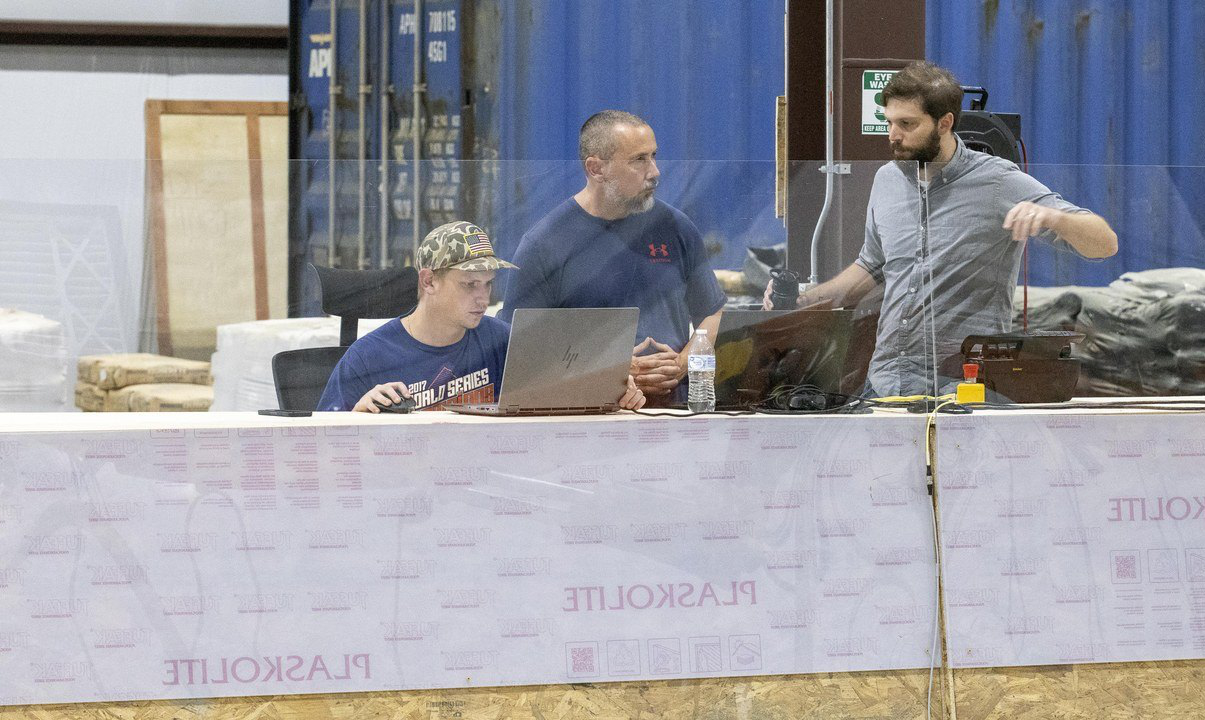 Three AUTOCON engineers working at a bench behind a safety screen in the shop, laptops and controls in front of them, a robotic arm and print nozzle reaching in from the right